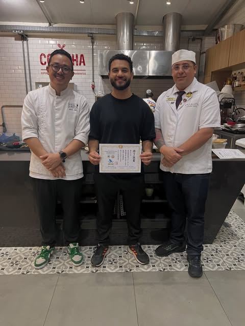 Thiago Cardoso segurando um certificado ao lado de dois chefs de cozinha japonesa